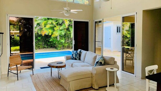 A spacious living area with a sofa, a small round table, and a chair. There is a large window providing a view of a pool and greenery outside. - Come have Fun in DR! (Sosúa)
