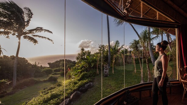 Ein Zimmer mit großem Fenster und Blick auf eine tropische Landschaft mit Palmen und Meer.