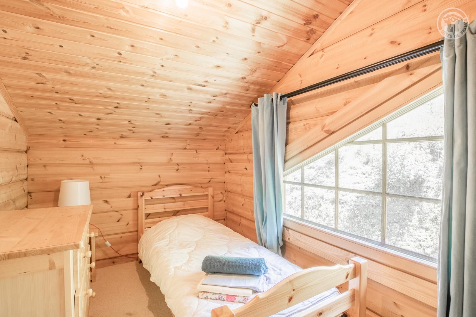 A wooden cabin room with a bed, a small desk, and a window with curtains.