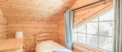 A wooden cabin room with a bed, a small desk, and a window with curtains.