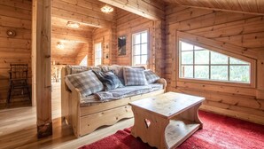 A cozy wooden cabin interior with a sofa, a small table, and a window offering a view of the forest.