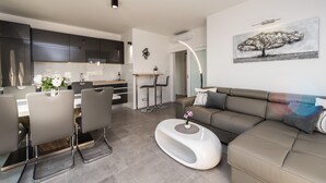 A modern living room with a sofa, a coffee table, and a dining area with chairs and a table. There is a kitchen in the background with dark cabinets and a white countertop. - Villa Magnifica Penthouse with Jacuzzi (Novalja)
