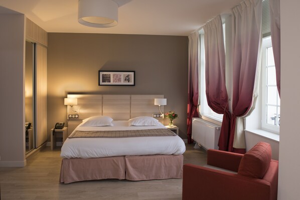 Habitación de hotel con una cama grande, mesitas de noche, una silla, ventana con cortinas y una obra de arte colgada en la pared.