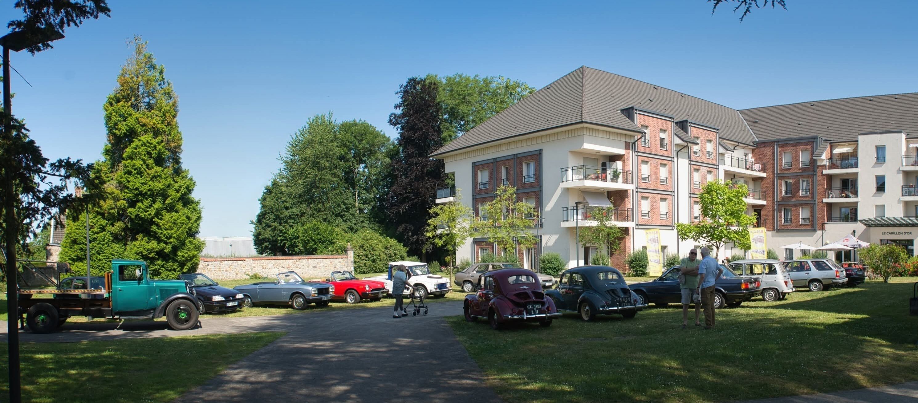 Un espace de stationnement où sont garées différentes voitures, un chemin menant à un bâtiment à plusieurs étages et une zone herbeuse avec des arbres.