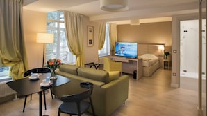 A modern hotel room with a sofa, a dining table, a TV, and a small desk area. - Manoir Le Carillon dOr (Maubeuge)