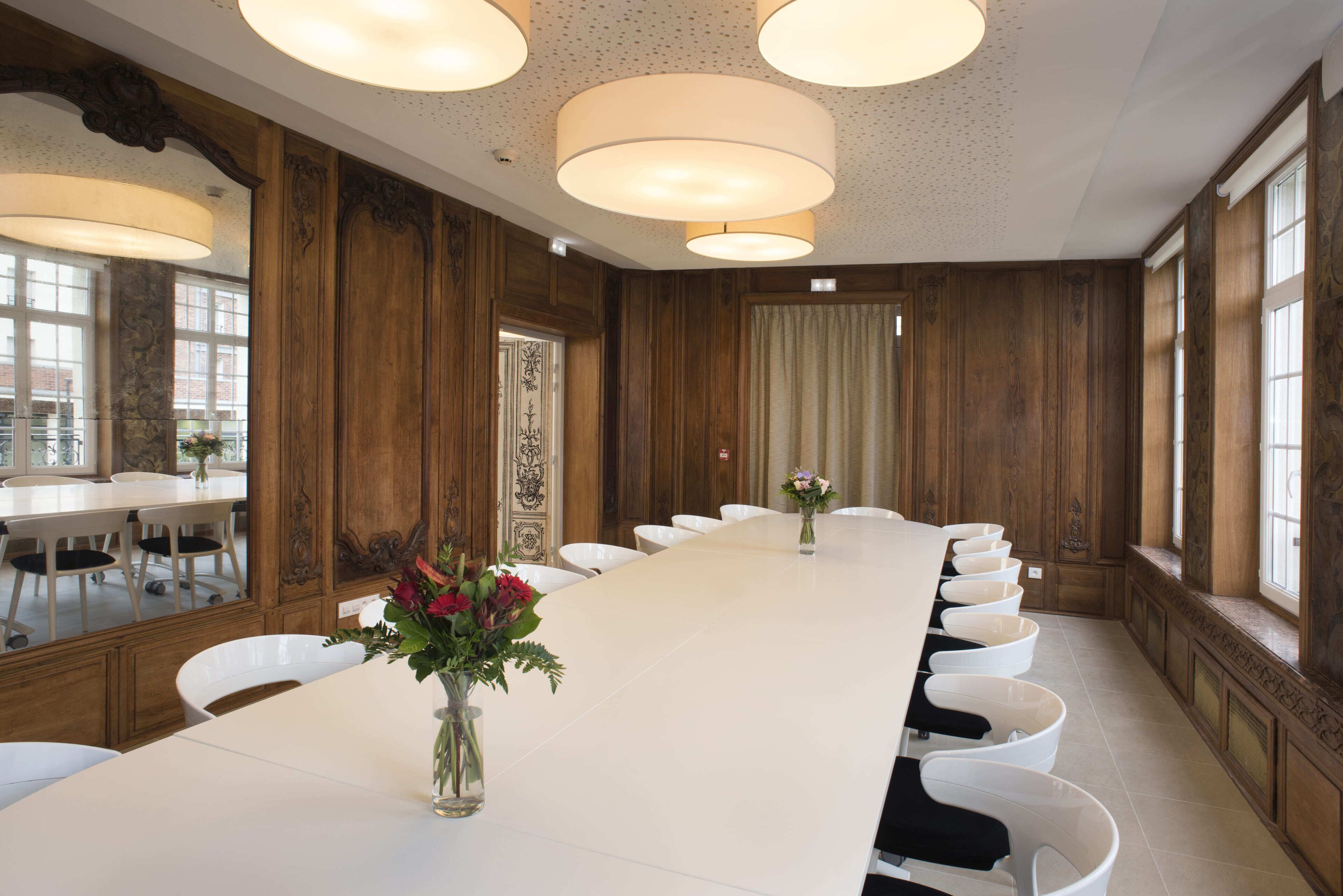 Une salle de conférence avec une longue table, des chaises blanches et un vase rempli de fleurs.