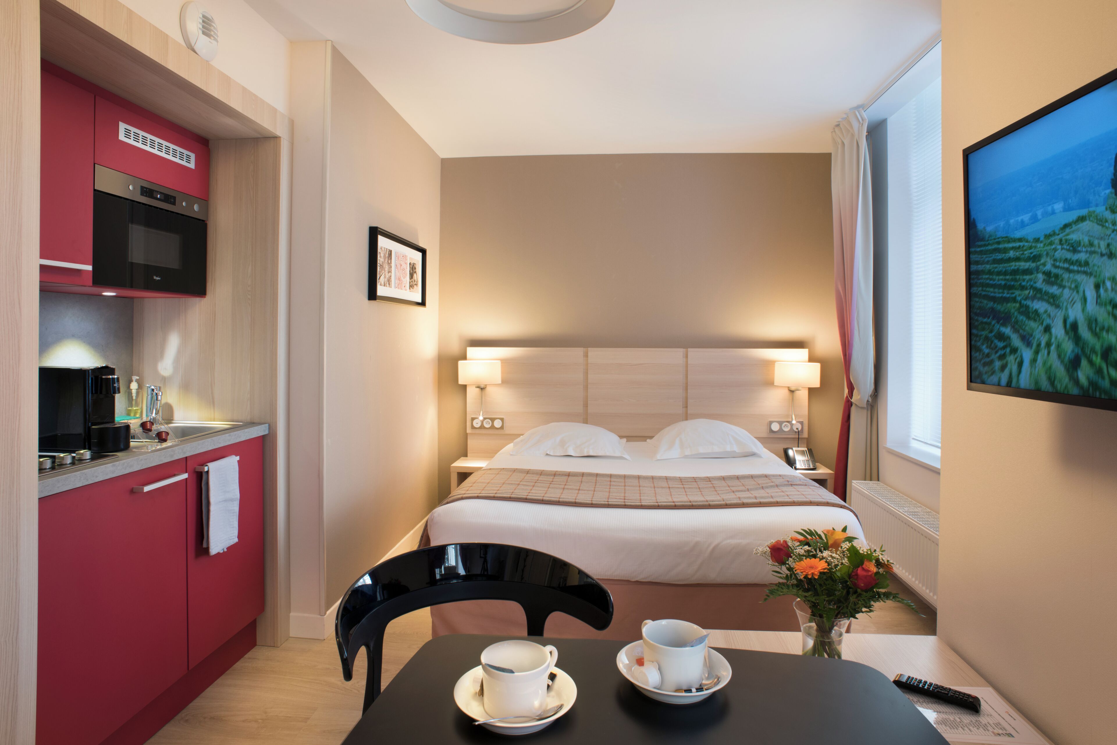Une chambre d’hôtel compacte comprenant un coin cuisine, un lit avec une tête de lit, des lampes de chevet, une table avec une tasse et une soucoupe, et un vase avec des fleurs.