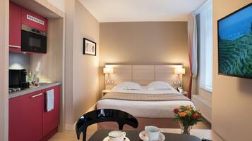 Une chambre d’hôtel compacte comprenant un coin cuisine, un lit avec une tête de lit, des lampes de chevet, une table avec une tasse et une soucoupe, et un vase avec des fleurs.