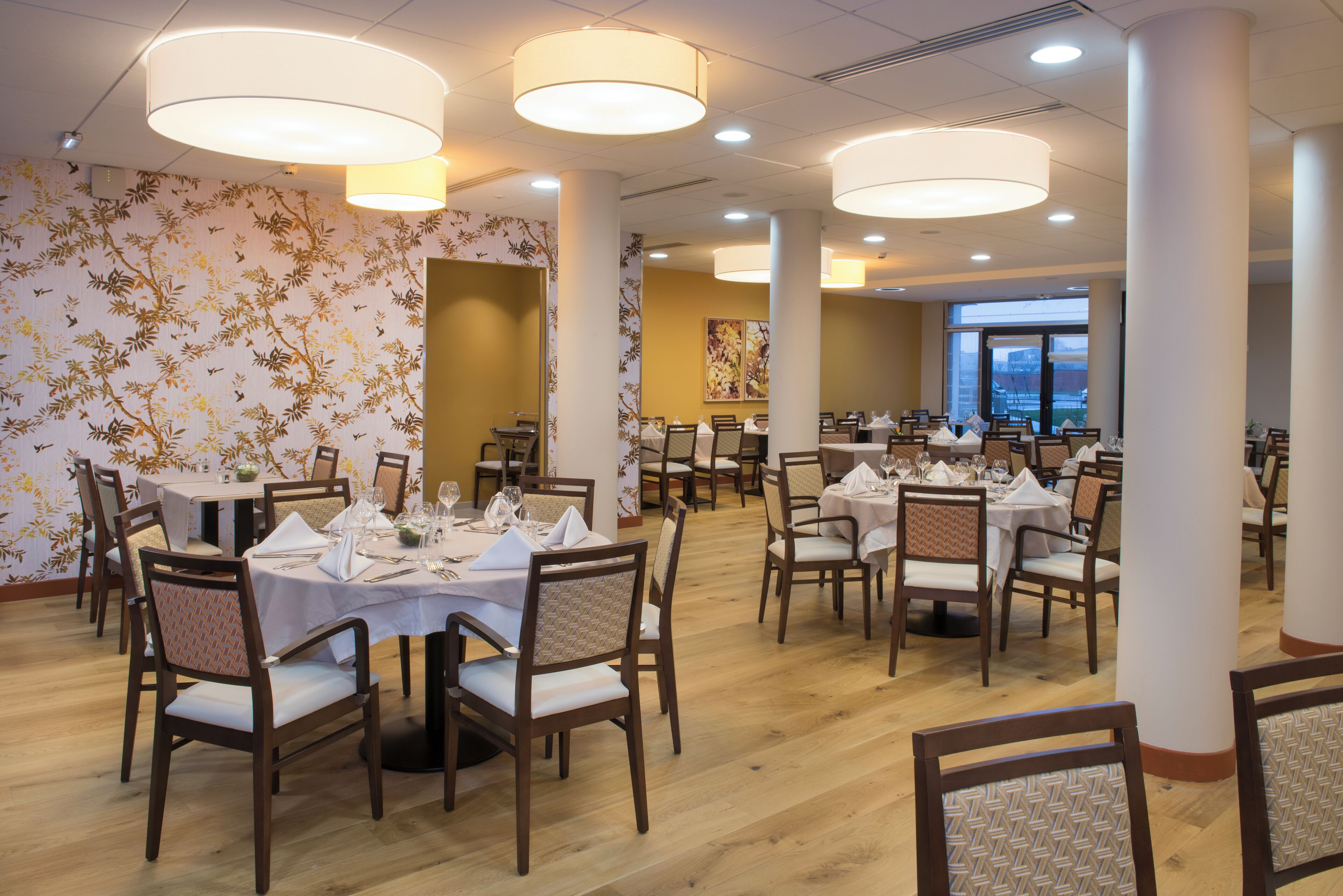 A dining area with multiple tables set for guests, featuring a floral wallpaper design and modern lighting fixtures.