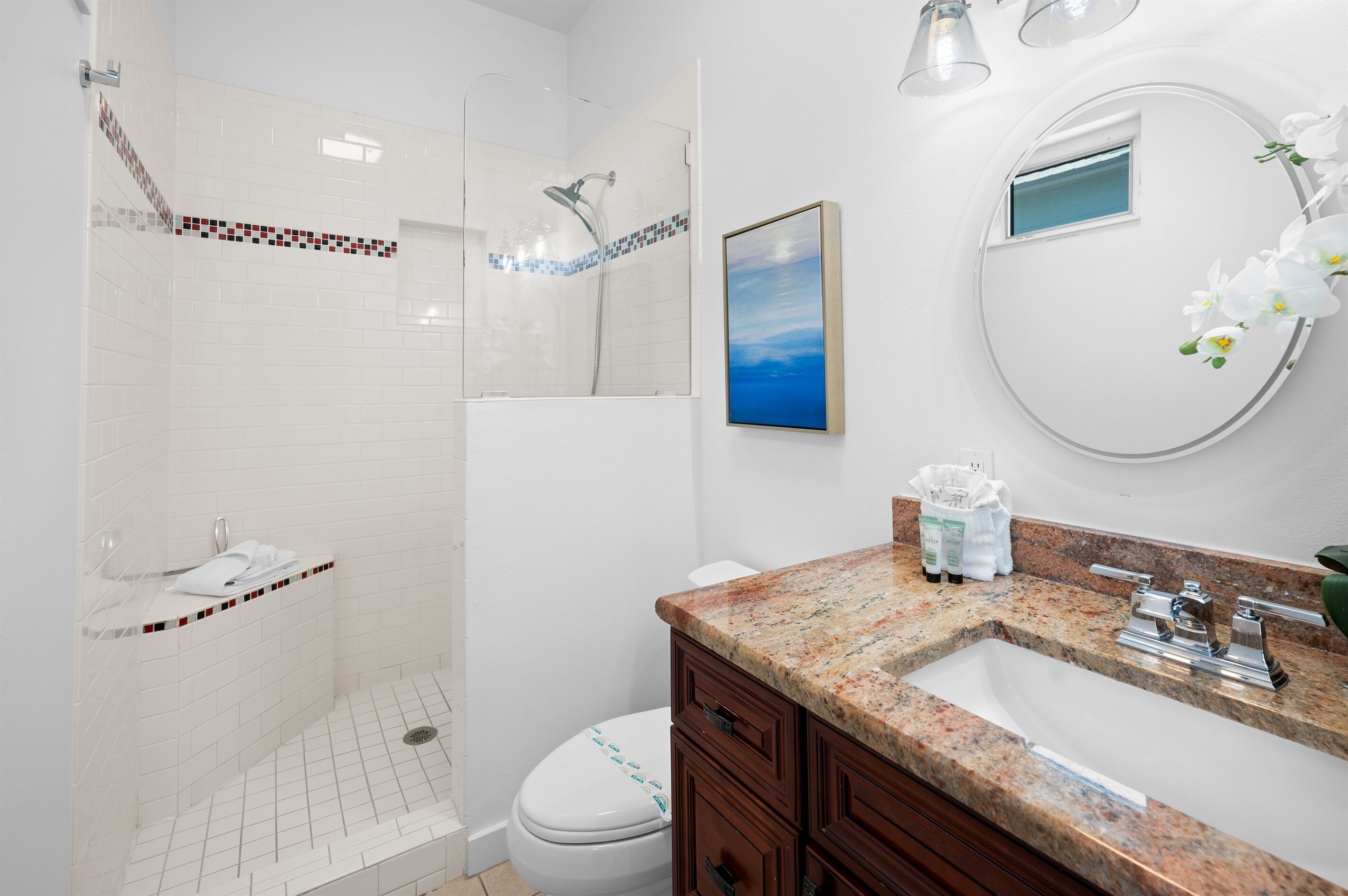 A modern bathroom with a glass-enclosed shower, a white toilet, a granite countertop sink, and a framed picture on the wall.