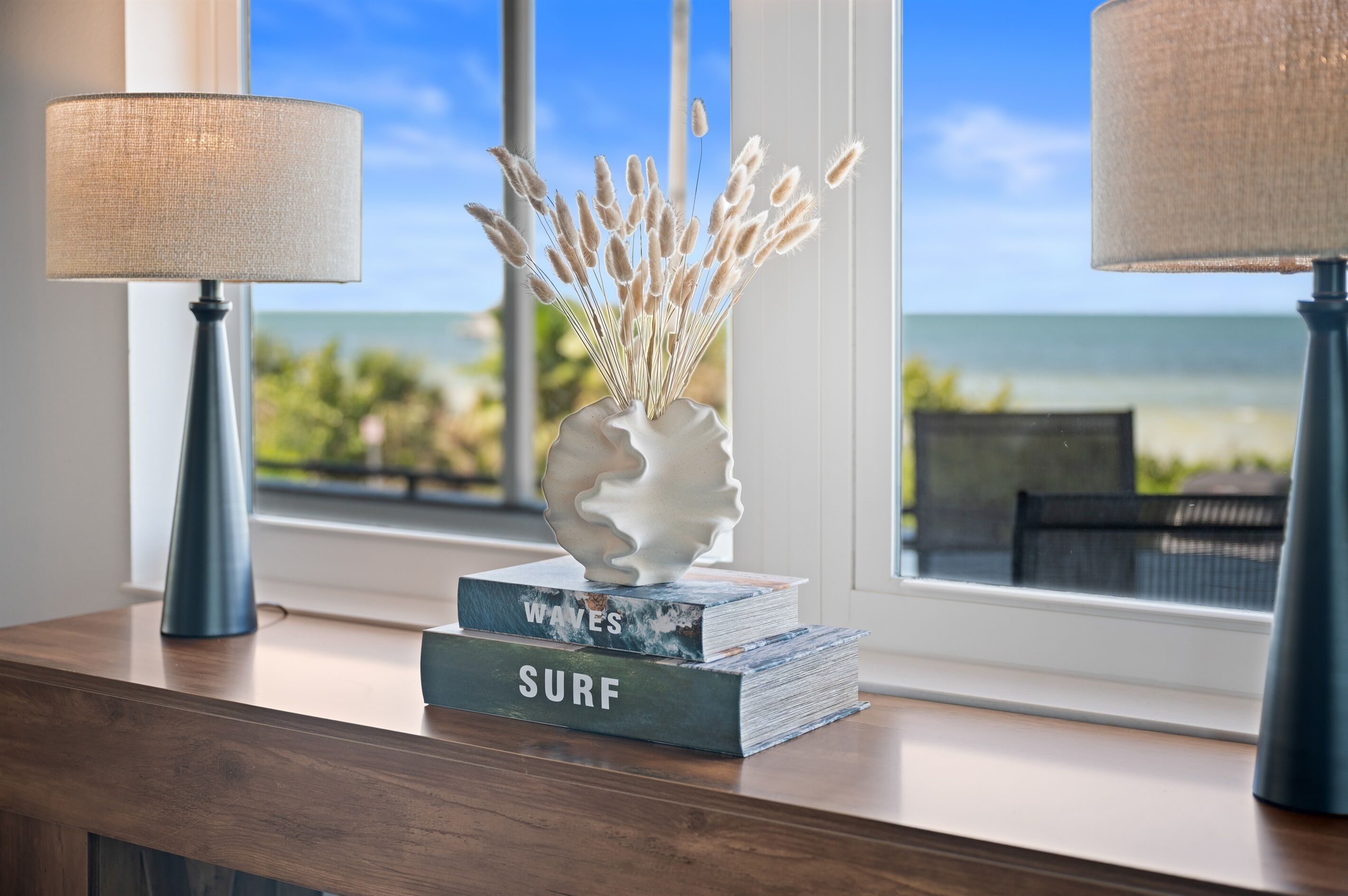 A wooden table with two lamps, a vase with dried grass, and two books titled "WAVES" and "SURF" in front of a window with a view of the ocean.