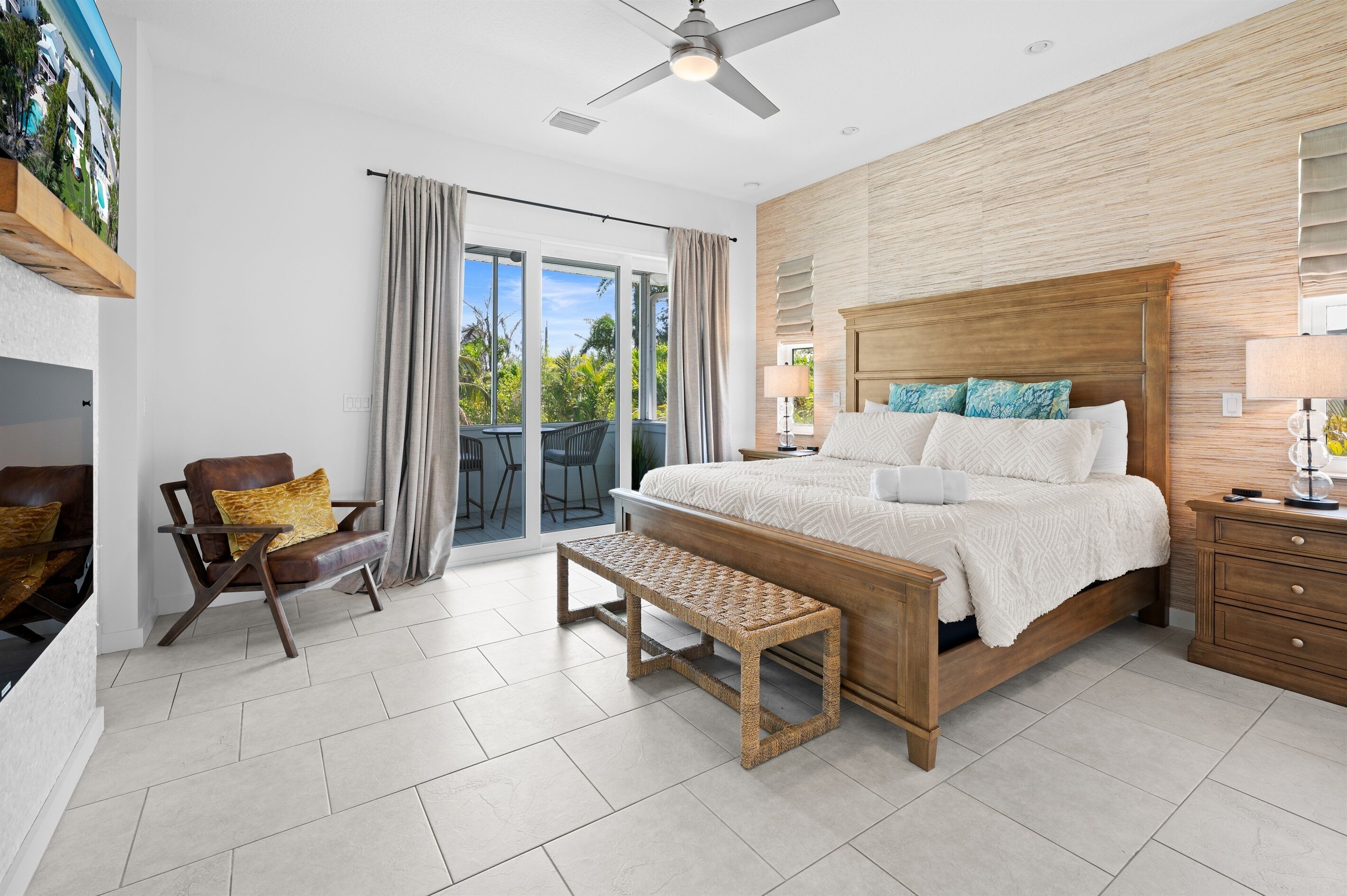 A modern bedroom with a large bed, a bench, a chair, and a view of the outdoors through a sliding door.