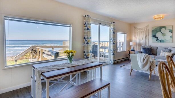 A spacious living room with a wooden floor, a dining area with a bench, and large windows offering a view of the beach and ocean. - Ocean Front Duplex with Views! Dog Friendly! Free Wifi! (South Beach)