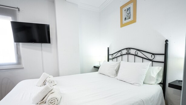 A neatly made bed with white linens and pillows, a black headboard, a flat-screen TV mounted on the wall, and a framed picture above the bed.