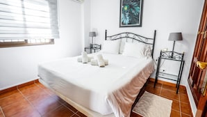 A neatly made bed with white linens, two bedside tables with lamps, a framed tropical leaf artwork on the wall, and a window with blinds. - Marbella Village | Patio View Terrace (Marbella)