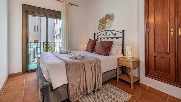 A bedroom with a bed, a nightstand, a wooden door, and a window with a view of buildings. - Marbella Village  | Appartment with Terrace (Marbella)