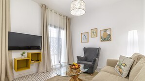 A living room with a flat-screen TV, a grey armchair, a beige sofa, a glass coffee table, and a small plant on a yellow shelf. - Casa Castillo | Ground Floor Unit (Marbella)