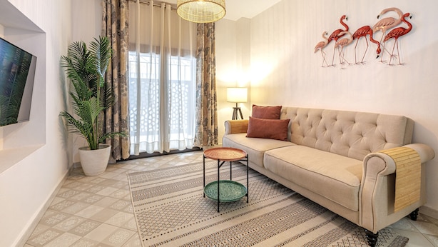 Una sala de estar con sofá, televisor, lámpara, planta en maceta, alfombra estampada y un cuadro de flamencos colgado en la pared.