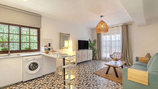 A compact living space with a kitchen, dining area, and seating area, featuring a patterned floor, a washing machine, a television, and a large window.