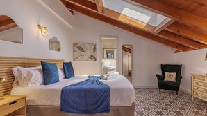 Ein Schlafzimmer mit schrägem Dach, einem Bett mit blauer Decke, einem Holz-Kopfteil, einem Stuhl, einer Kommode und einem gemusterten Boden.