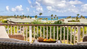 Parveke, jossa on rottinkihuonekaluja ja hedelmäkulho, ja josta avautuu näkymä palmupuiden reunustamalle rannalle ja kirkkaalle taivaalle.