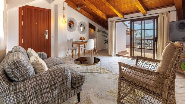 Ein gemütliches Wohnzimmer mit Holzbalkendecke, einer Couch, einem Korbstuhl, einem Esstisch mit Stühlen und einem großen Fenster mit Aussicht.