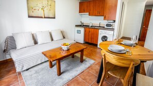 A compact living space with a sofa, a wooden dining table set for two, a kitchen area with wooden cabinets, and a painting on the wall.