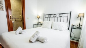 A neatly made bed with white linens and two rolled towels, a metal-framed headboard, bedside lamps, and a bathroom visible through an open door.
