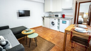 En kompakt opholdsstue med sofa, et lille spisebord, et tv ophængt på væggen, en vaskemaskine og en mikrobølgeovn.