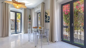 A modern dining area with a glass table and chairs, a hanging light fixture, and a view through a glass door to an outdoor balcony with plants.