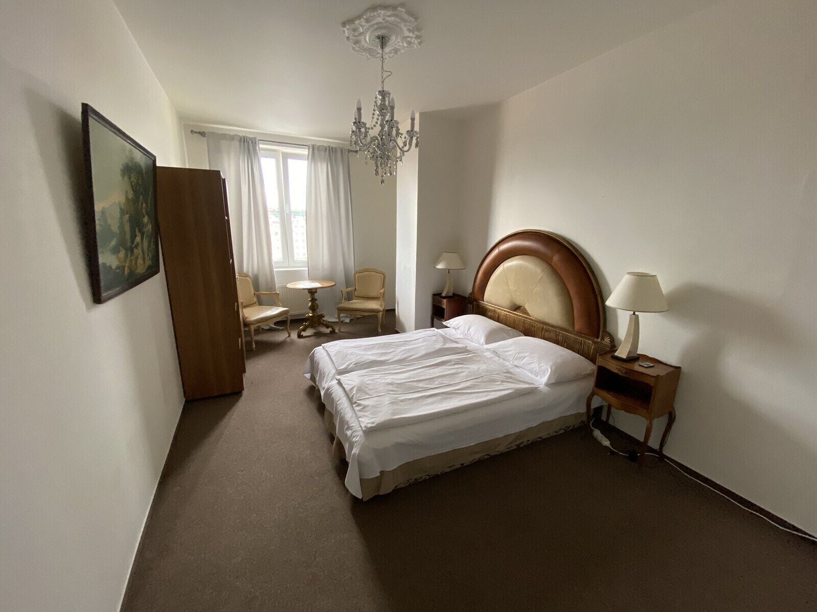 A hotel room with a large bed, two bedside lamps, a chandelier, a desk with a chair, and a painting on the wall.