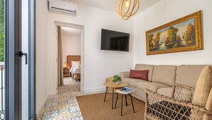 A living room with a sofa, a wicker chair, a small table with a plant, a painting on the wall, and a TV. - Casa Serena  | Family Apartment (Marbella)