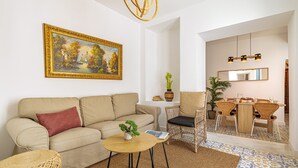 A living room with a sofa, a coffee table, a painting, and a dining area with a table and chairs. - Casa Serena  | Family Apartment (Marbella)