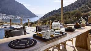 En terrasse med et træbord, stole, en bakke med citroner, glas og stearinlys, med udsigt over et kystlandskab med bjerge og en landsby.
