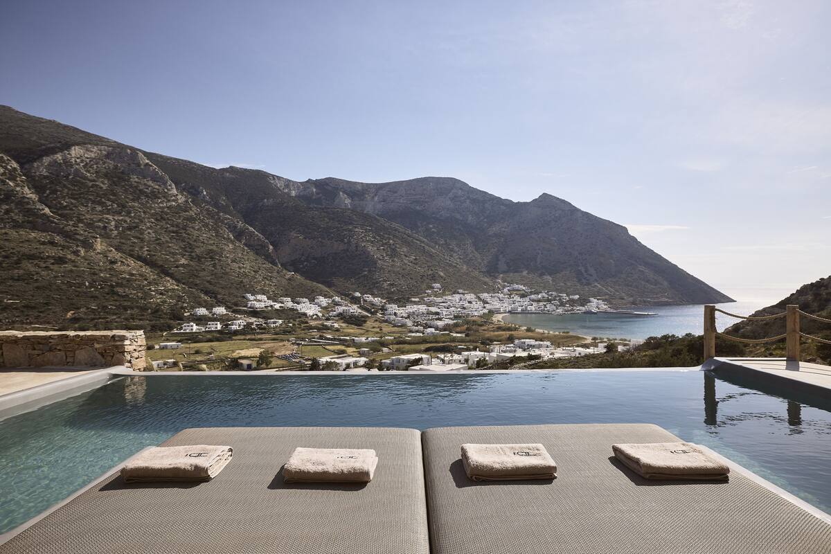 Piscina infinita con sillones de descanso y vistas a un pueblo costero y montañas.