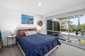 A bedroom with a large bed, a painting, a round wooden shelf, a desk, and a view of the outdoors.