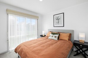 A bedroom with a bed, a nightstand, a bedside table, a framed picture on the wall, and a window with a curtain. - Gumnut House (Anglesea)