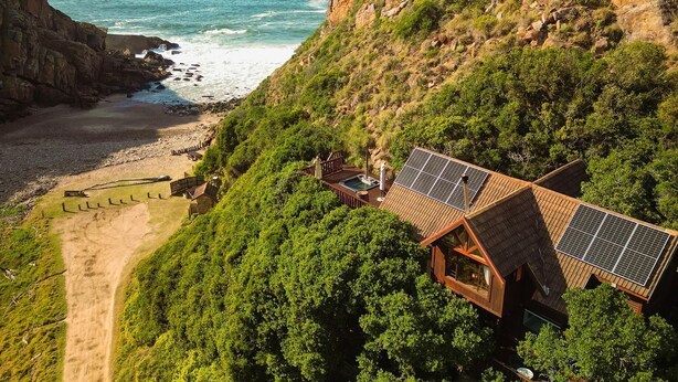 Una cabaña de madera con paneles solares, situada en una ladera con vistas a una playa y el océano.