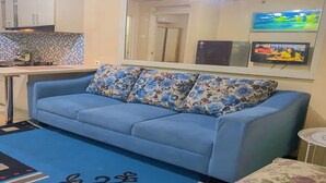 A living room with a blue sofa, a kitchen in the background, and a large mirror reflecting the room.