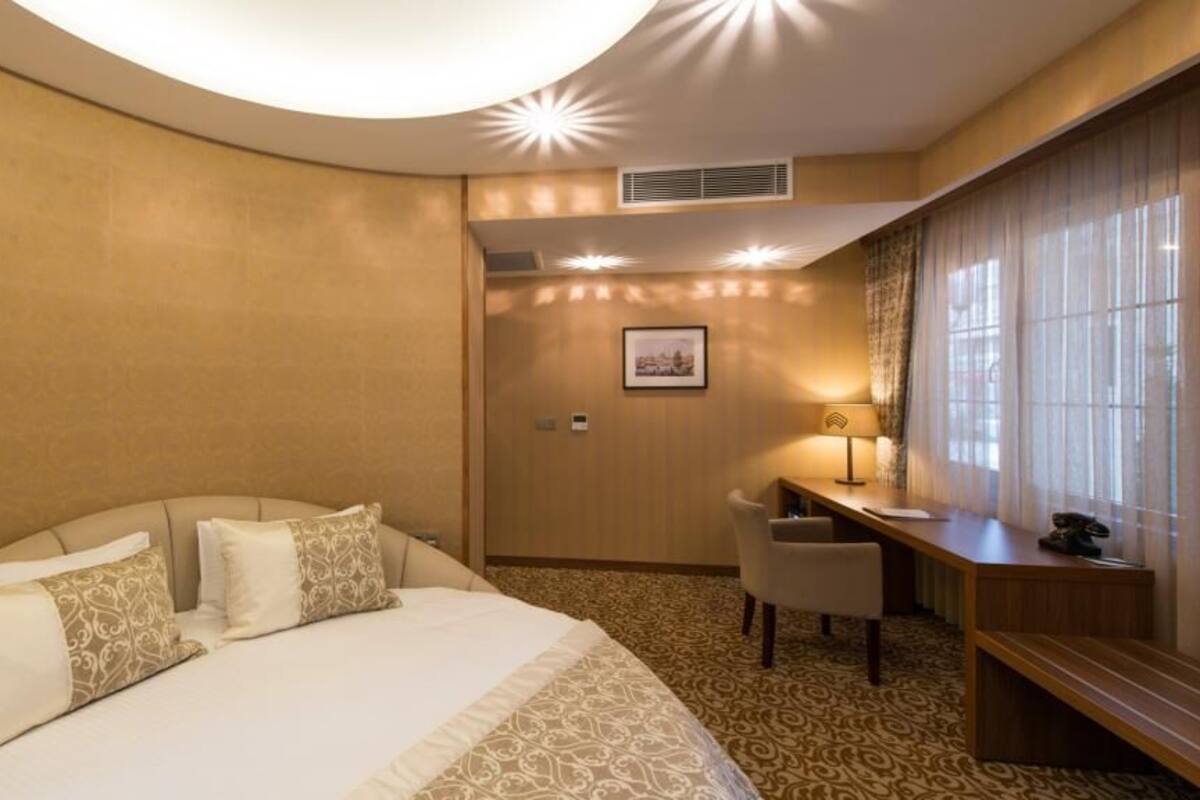 A hotel room with a large bed, a desk, and a chair. There is a picture on the wall and a lamp on the desk.