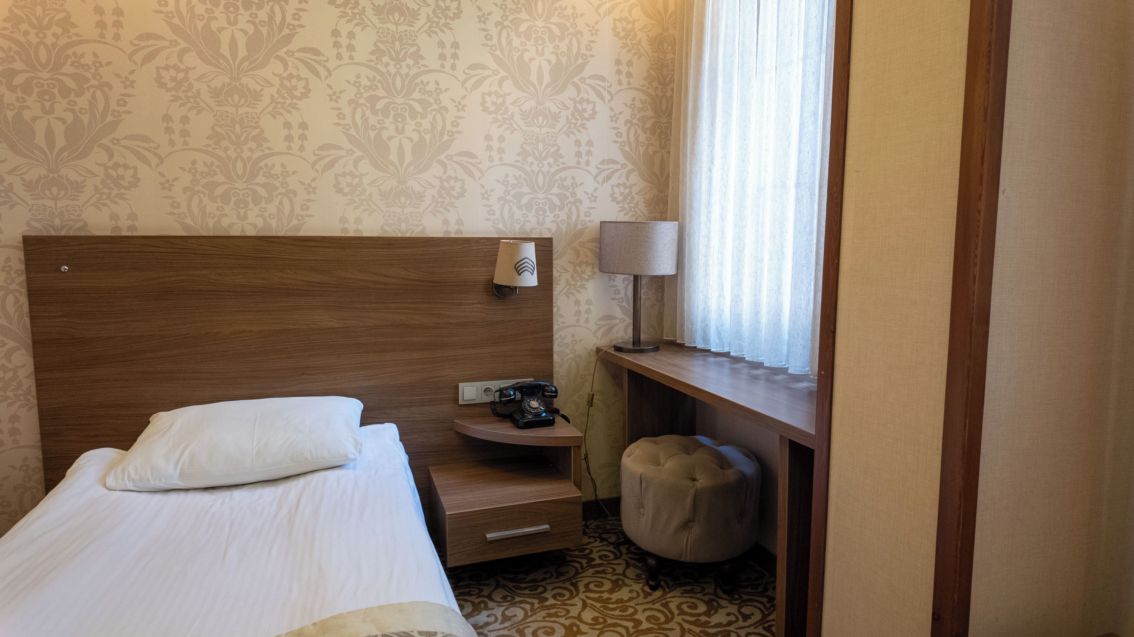 A hotel room with a wooden headboard, a single bed with white bedding, a bedside table with a lamp and a telephone, a small ottoman, and a window with sheer curtains.