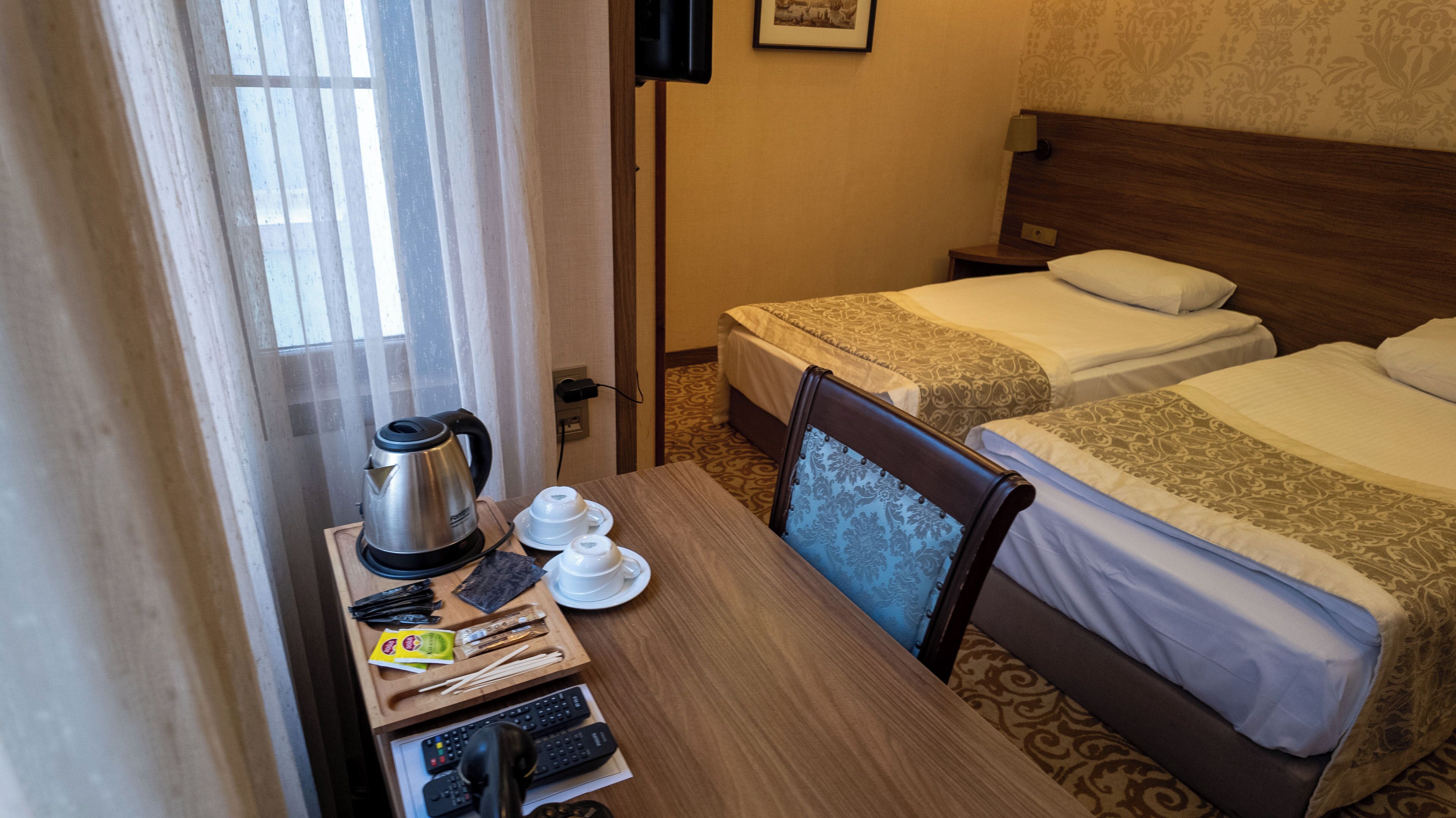 A hotel room with two beds, a wooden table with a kettle, cups, and remote controls.