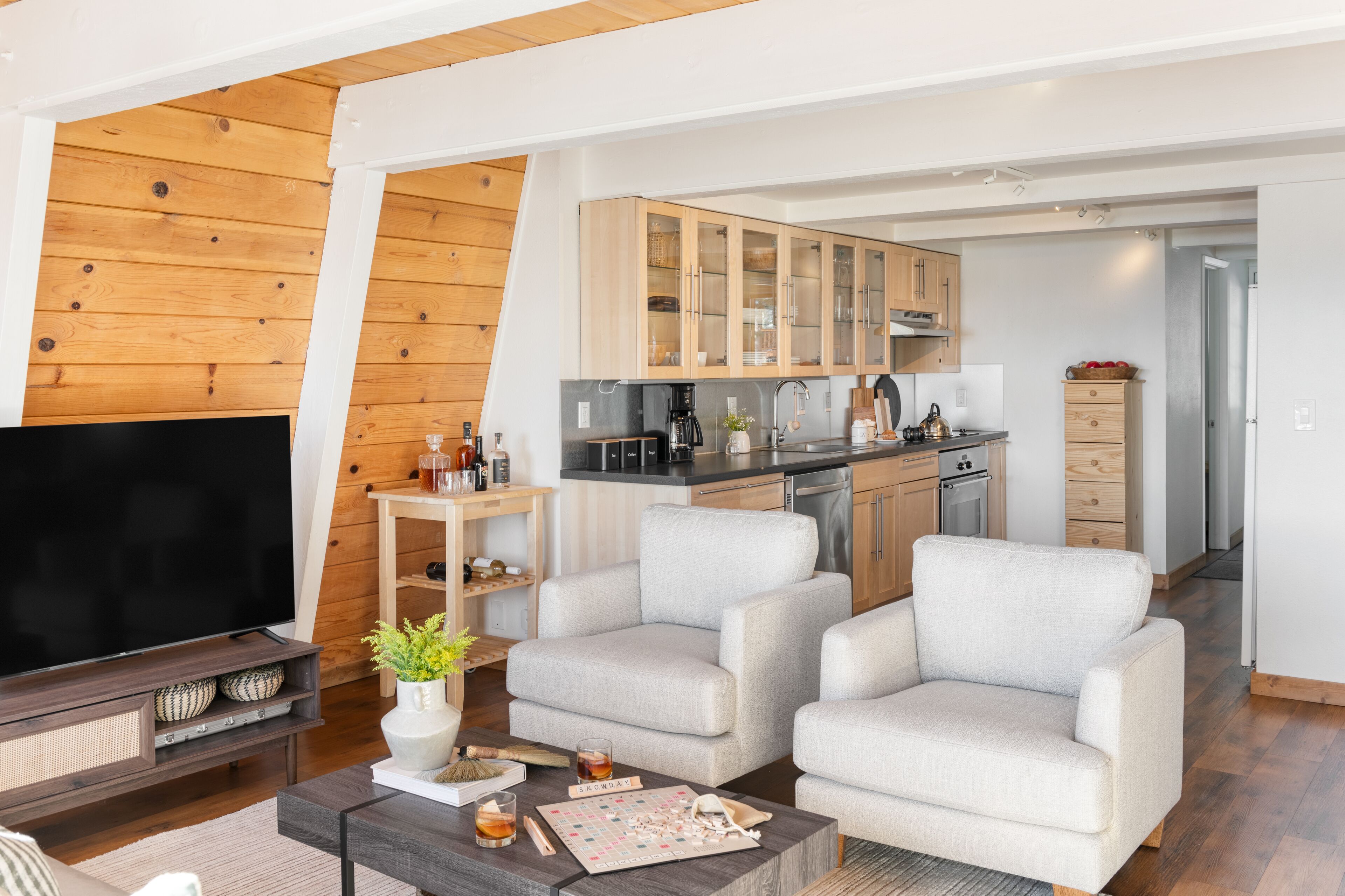 Un salon moderne doté d’un téléviseur à écran plat, de deux fauteuils, d’une table basse avec des jeux de société, et d’une cuisine en arrière-plan.