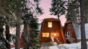 Een houten chalet met een trap naar de ingang, omringd door een met sneeuw bedekte omgeving en hoge bomen.