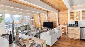 Een moderne woonkamer met een bank, salontafel en televisie. Er is een keukenhoek met kastjes en een vaatwasser. Grote ramen bieden uitzicht op het omliggende landschap.