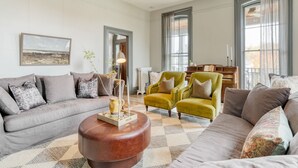 Un salon spacieux avec un canapé gris, un fauteuil jaune et un pouf marron. Une grande fenêtre avec des rideaux, un tableau au mur et un vase avec des bougies sur le pouf complètent la pièce.