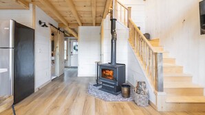 A spacious room with a wooden floor, a black wood stove, a wooden staircase, and a modern kitchen with a stainless steel refrigerator.