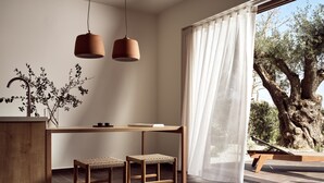 Cozinha moderna com mesa de madeira, dois taburetes de vime, luminárias pendentes e vista para uma oliveira através de cortinas translúcidas.