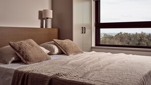Quarto moderno com uma cama grande, cabeceira, mesa de cabeceira e janela com vista para árvores.