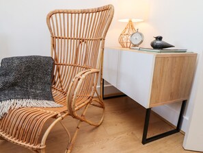 Une chaise en osier, avec une assise et un dossier tissés, est placée à côté d’une armoire blanche dotée d’un socle en bois. Une lampe avec un abat-jour sphérique et une figurine d’oiseau se trouvent sur l’armoire.